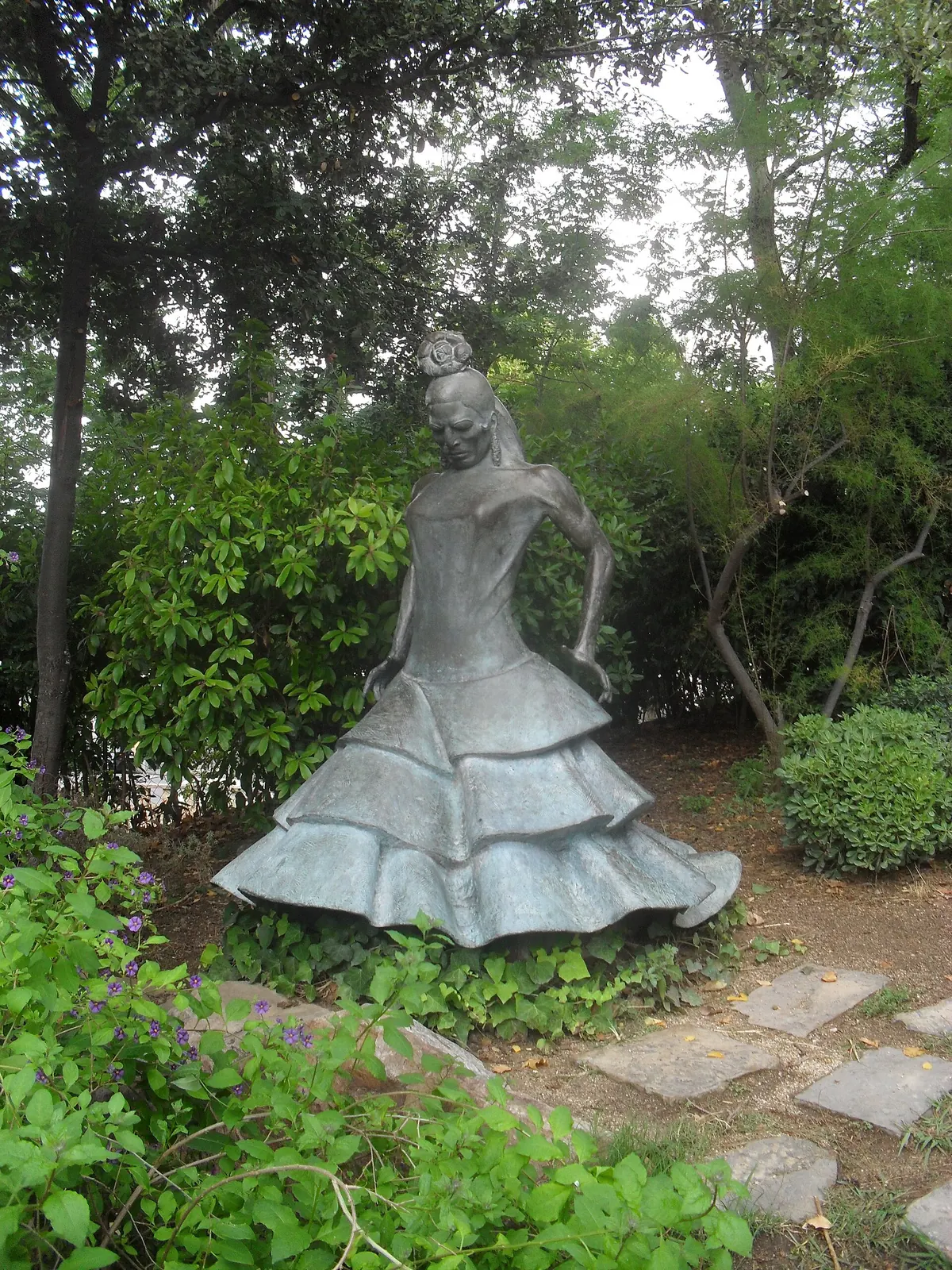 A grey metal sculpture of a flamenco dancer in a flounced dress among green shrubs
