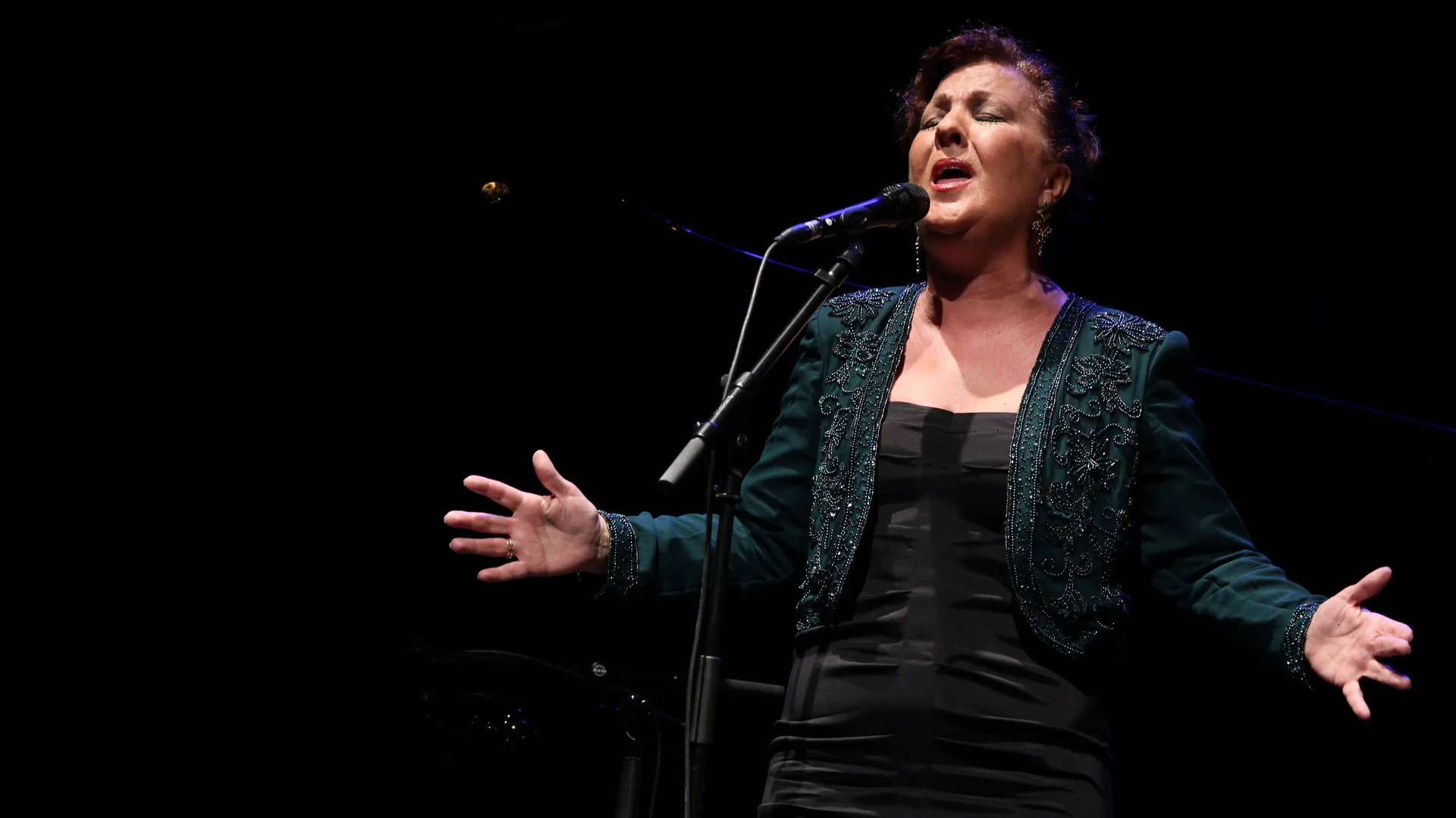A flamenco singer with open hands, mid-song at a microphone on a dark stage, in a green beaded jacket