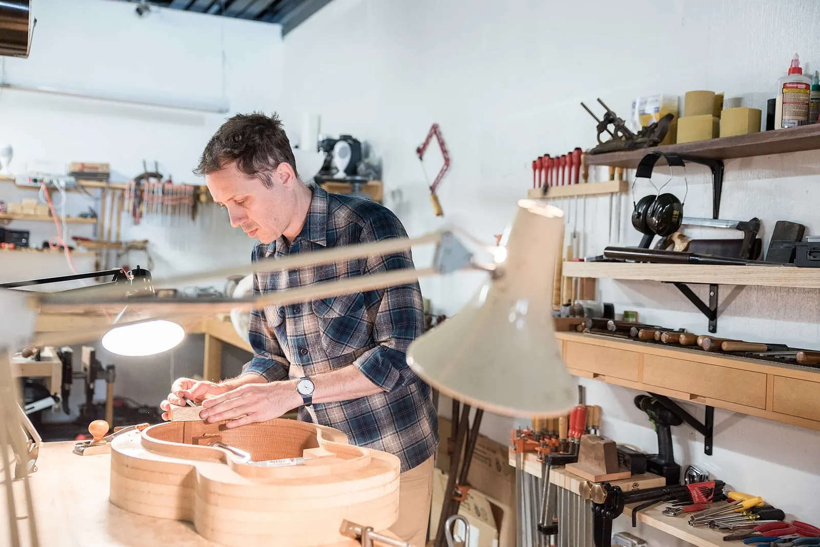 A guitar maker shaping the curved body of an instrument at a workbench, with hand tools on the shelves behind him